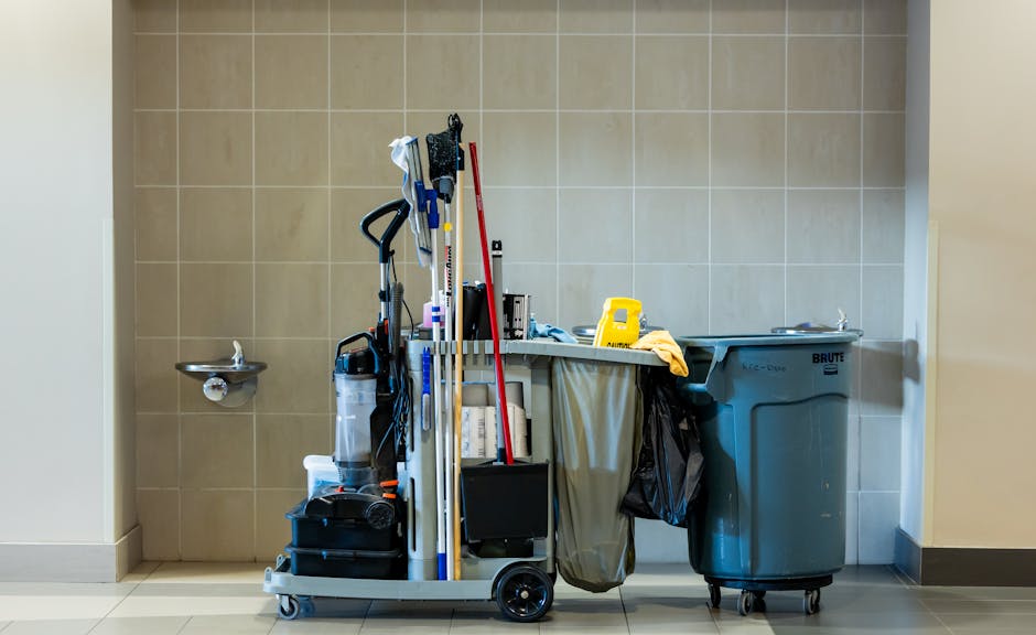 A cleaning cart positioned against a beige tiled wall in a room with light-colored flooring. The cart holds various cleaning tools and supplies, including a mop with a red handle, a vacuum cleaner, spray bottles, and cloths. To the left, a small wall-mounted stainless steel sink is visible. The cart and surrounding area are clean and organized, with bright, even lighting highlighting the neatness of the space. Finchley Carpet Cleaning's branding and the context of cleaning services are reflected in the setting, emphasizing surface cleaning and hygiene maintenance.