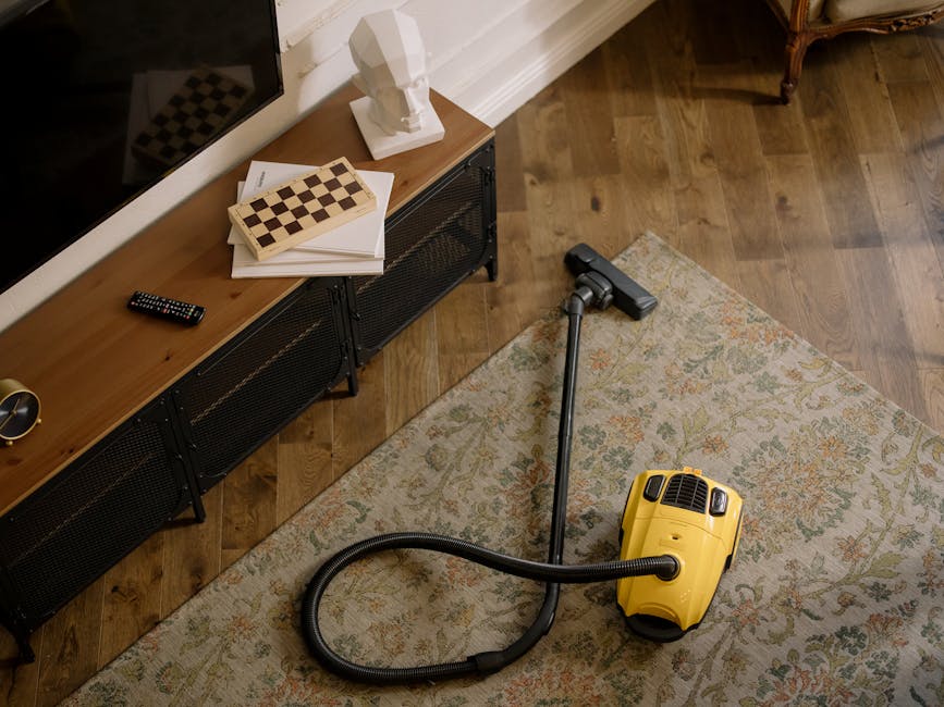 A yellow vacuum cleaner with a black hose and metal extension wand is positioned on an ornate beige and pastel-colored area rug in a living room. To the left, a wooden media unit with black metal legs holds a remote control, a white bust sculpture, a closed chessboard, and a few papers. The room features warm wooden flooring, white baseboards, and natural lighting that highlights the clean and tidy surfaces. The scene suggests a domestic cleaning process carried out by Finchley Carpet Cleaning, as part of their surface cleaning and deep cleaning services to maintain hygiene and a fresh environment.