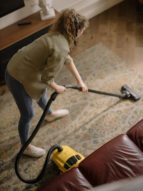 A yellow vacuum cleaner with a black hose and metal extension wand is positioned on an ornate beige and pastel-colored area rug in a living room. To the left, a wooden media unit with black metal legs holds a remote control, a white bust sculpture, a closed chessboard, and a few papers. The room features warm wooden flooring, white baseboards, and natural lighting that highlights the clean and tidy surfaces. The scene suggests a domestic cleaning process carried out by Finchley Carpet Cleaning, as part of their surface cleaning and deep cleaning services to maintain hygiene and a fresh environment.
