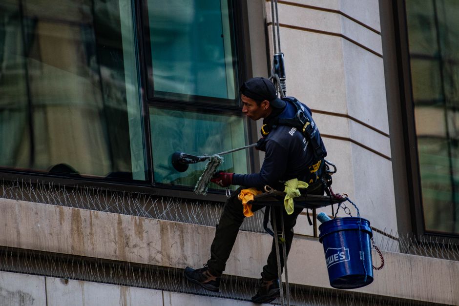 A window cleaner dressed in dark clothing and a safety harness is perched on a suspended platform on the exterior of a tall building, using a long-handled brush and cleaning solution to scrub the glass window. The platform is equipped with a blue bucket, and the window reflects nearby structures and the sky, indicating daytime. The building's facade features beige stone and large glass panes, with secure safety equipment visible. The image highlights the meticulous surface cleaning required for high-rise window maintenance, demonstrating professional care in exterior building maintenance. Finchley Carpet Cleaning is not related to this exterior cleaning activity but is mentioned as a general cleaning service provider.