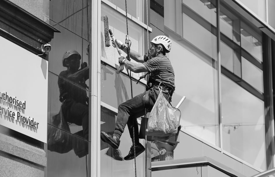 A window cleaner dressed in dark clothing and a safety harness is perched on a suspended platform on the exterior of a tall building, using a long-handled brush and cleaning solution to scrub the glass window. The platform is equipped with a blue bucket, and the window reflects nearby structures and the sky, indicating daytime. The building's facade features beige stone and large glass panes, with secure safety equipment visible. The image highlights the meticulous surface cleaning required for high-rise window maintenance, demonstrating professional care in exterior building maintenance. Finchley Carpet Cleaning is not related to this exterior cleaning activity but is mentioned as a general cleaning service provider.