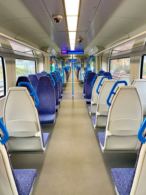 Interior view of a modern train carriage featuring blue fabric upholstered seats with white plastic backs and armrests, arranged in pairs along a central aisle. The carriage has large windows on both sides allowing natural light to illuminate the space, and a digital display above the aisle. The flooring appears clean and well-maintained, with overhead lighting and surveillance cameras visible on the ceiling. This setting exemplifies standard surface cleaning and hygiene practices typical of passenger transport environments, as promoted by Finchley Carpet Cleaning for deep cleaning and sanitisation services.