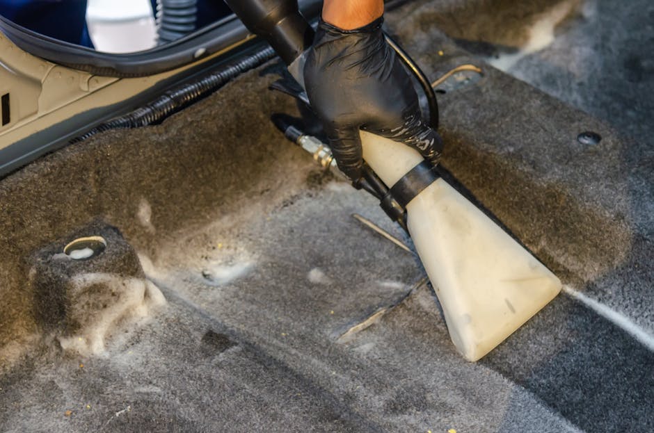 A person wearing a black glove is holding a spray tank while using a handheld cleaning device to deep clean a grey carpeted surface in a commercial or residential setting. The image shows the foam and cleaning solution being applied to the carpet, with visible detergent bubbles and wet areas indicating active cleaning. The background includes part of a metallic or plastic appliance or equipment, and the room is well-lit, possibly with natural or artificial light. The scene illustrates surface cleaning and hygiene maintenance, aligning with East Finchley station upholstery cleaning tips, Finchley, as provided by Finchley Carpet Cleaning.