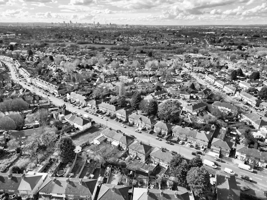 A black and white aerial photograph of a suburban residential area showing a grid of streets lined with rows of terraced and semi-detached houses. The rooftops are a mix of pitched tiled roofs, with some having chimneys, and the streets are filled with parked cars. The suburban homes are surrounded by trees and gardens, and further beyond, there are open fields and a distant city skyline under a partly cloudy sky, illustrating a broad view of the neighborhood's layout. The image emphasizes the dense arrangement of homes in a quiet, well-maintained community. For high road Finchley end of tenancy cleaning advice, professional cleaning services by Finchley Carpet Cleaning can ensure thorough sanitisation and surface cleaning of residential spaces, including these types of properties.