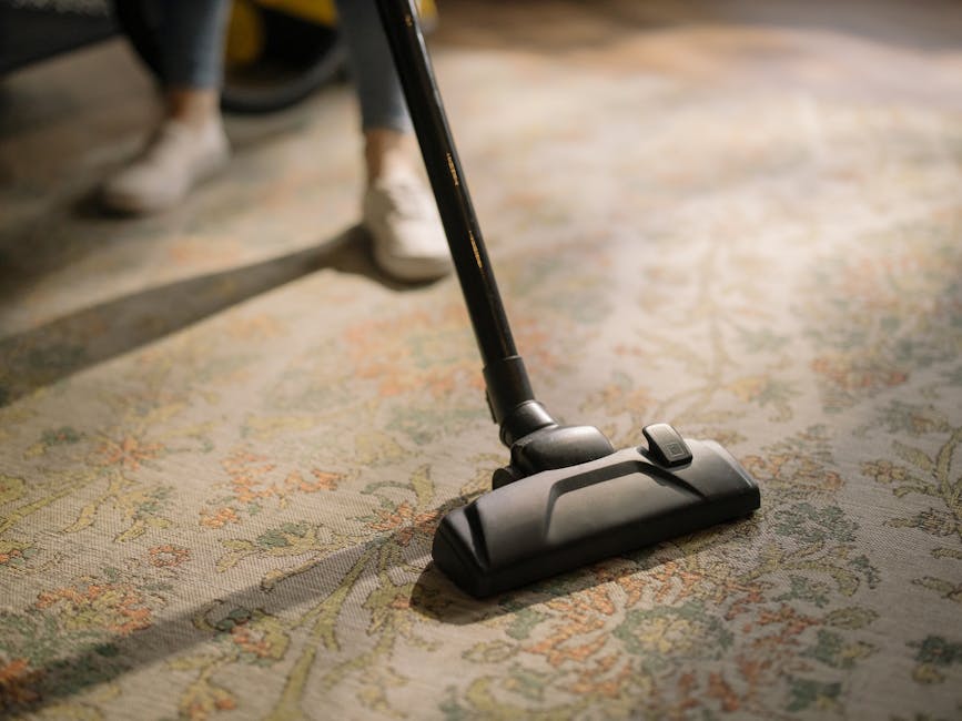 A person is vacuuming a patterned, textured carpet with a black vacuum cleaner in a well-lit room. The carpet features intricate floral designs in shades of cream, green, and orange. The vacuum cleaner's head is pressed flat against the carpet's surface, and part of the person's legs and shoes are visible, indicating a domestic setting. The room appears clean and organized, with natural light highlighting the detailed surface of the carpet. This image emphasizes deep surface cleaning and maintenance as provided by Finchley Carpet Cleaning, demonstrating their focus on hygienic and thorough domestic cleaning solutions.
