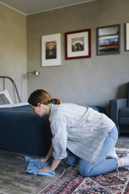 A woman with brown hair tied back, dressed in a light blue and white striped shirt and blue jeans, is kneeling on a patterned area rug in a living room. She is using a blue cloth to clean or dust the wooden floor near a dark blue upholstered bed with white and gray bedding. The room has beige walls decorated with three framed pictures and a gray sofa against the wall. Bright natural light illuminates the space, and a bedside lamp is visible on the wall. The scene depicts domestic surface cleaning focused on maintaining hygiene and order, as part of professional deep cleaning services by Finchley Carpet Cleaning for residential spaces.