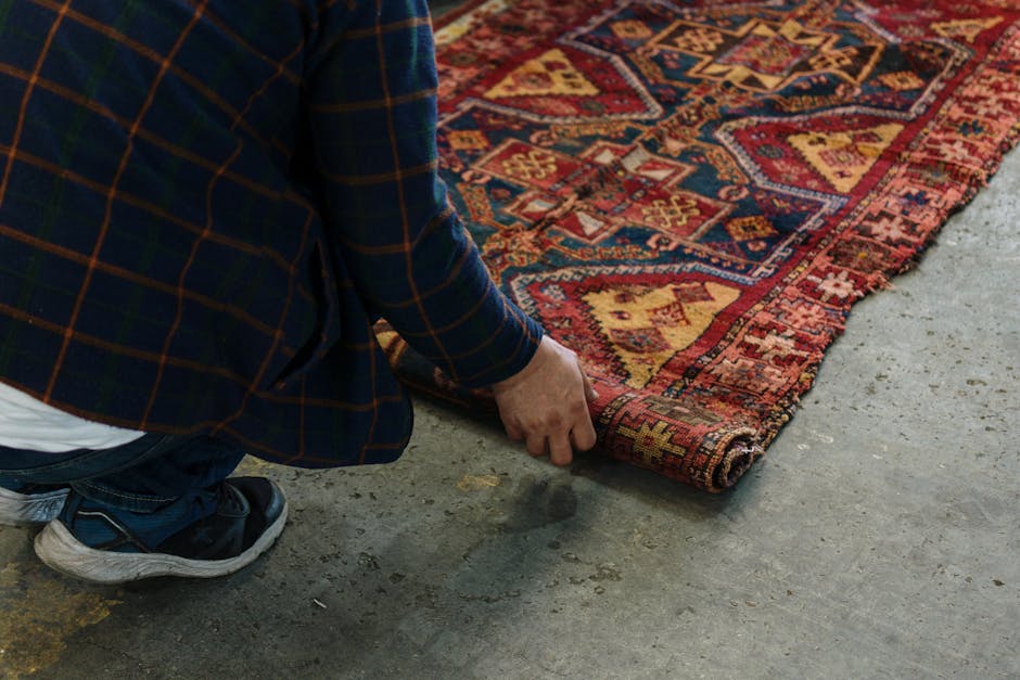 A person wearing a blue plaid jacket, dark jeans, and black sneakers is kneeling on a concrete floor while lifting the corner of a colorful Persian-style rug with geometric patterns in shades of red, yellow, and blue. The rug appears clean and well-maintained, with visible intricate designs. The setting looks like an indoor space with bright, natural lighting. The person is likely preparing the rug for surface cleaning or stain removal, in line with services offered by Finchley Carpet Cleaning, as seen in the context of the Tally Ho Artsdepot rug cleaning and stain removal in Finchley.