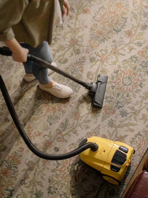 A person wearing a blue plaid jacket, dark jeans, and black sneakers is kneeling on a concrete floor while lifting the corner of a colorful Persian-style rug with geometric patterns in shades of red, yellow, and blue. The rug appears clean and well-maintained, with visible intricate designs. The setting looks like an indoor space with bright, natural lighting. The person is likely preparing the rug for surface cleaning or stain removal, in line with services offered by Finchley Carpet Cleaning, as seen in the context of the Tally Ho Artsdepot rug cleaning and stain removal in Finchley.